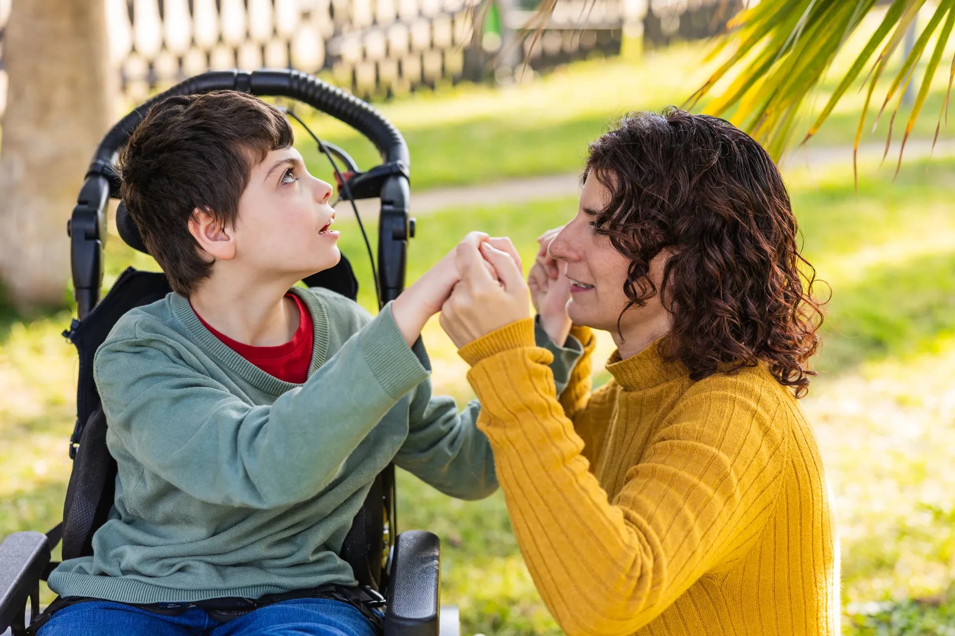 Una cuidadora agarra de las manos a un niño en silla de ruedas y le sonríe en un parque