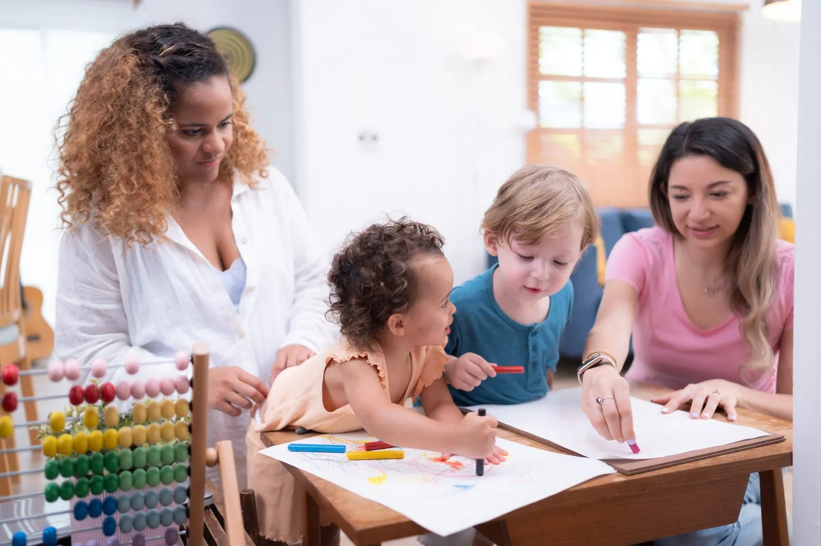 Dos adultas acompañan a unos niños que dibujan sentados a la mesa